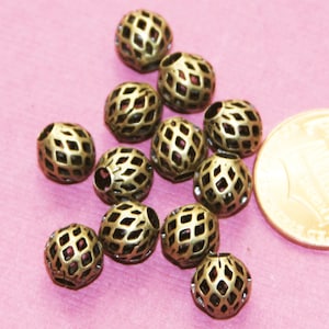 Puede incluir: Un conjunto de cuentas esféricas de color bronce con un diseño de celosía. Las cuentas están dispersas sobre una superficie rosa, con una moneda para comparar el tamaño. Los interiores huecos de las cuentas son visibles a través de las aberturas en forma de diamante.