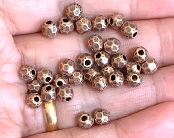 Könnte beinhalten: Nahaufnahme einer Hand, die eine Sammlung kleiner, kupferfarbener, gehämmerter Metallperlen hält. Die Perlen haben eine strukturierte, unregelmäßige Oberfläche und sind perfekt für die Schmuckherstellung.