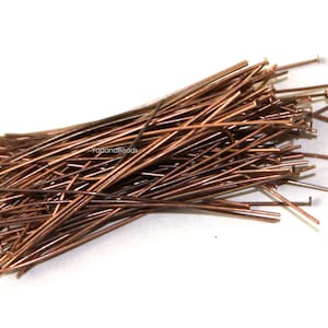 Peut inclure: Une grande collection d'épingles à tête en métal de couleur cuivre, chacune dotée d'une tige fine et droite avec une petite tête plate à une extrémité. Les épingles sont disposées en un tas lâche et superposé, avec le texte YadanaBeads.