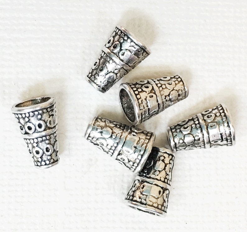 May include: Six silver metal cone-shaped bead caps with an intricate embossed design. The caps are scattered on a white surface.
