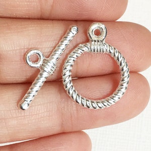 May include: Silver tone toggle clasp with a rope design. The clasp is made of two pieces, a round ring and a bar with a loop on one end.