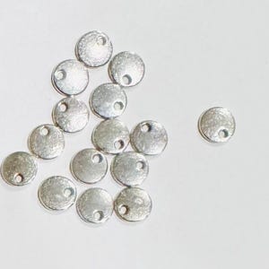 以下が含まれることがあります： 中央に小さな穴が開いた銀色の金属製の円がいくつか積み重ねられています。円は白い表面に緩く積み重ねられています。