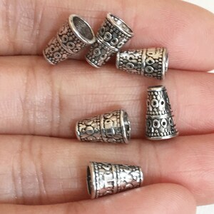 May include: Six silver metal cone-shaped bead caps with an intricate patterned design. The bead caps are approximately 1 cm in diameter.