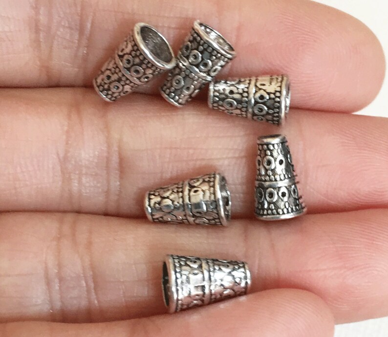 May include: Six silver metal cone-shaped bead caps with an intricate patterned design. The bead caps are approximately 1 cm in diameter.