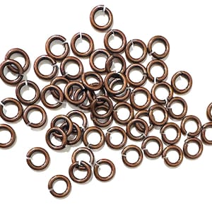 May include: A collection of numerous small, circular metal jump rings with an antique copper finish. Each individual ring features a single open cut along its circumference, allowing for easy connection and assembly in jewelry making projects.