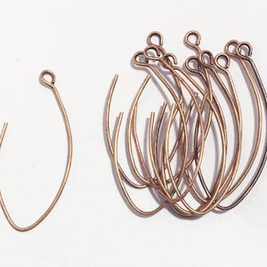 May include: A group of 10 gold-toned jump rings, each with a small loop at the top. The rings are arranged in a pile, with some overlapping. One ring is separated from the group and is lying flat.