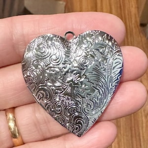 May include: A silver heart-shaped pendant with an ornate floral design. The pendant has a small loop at the top for attaching to a chain. The surface of the pendant is highly reflective, catching the light and creating a shimmering effect.
