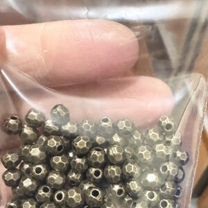 Pu&ograve; includere: Un sacchetto di perline in metallo dorato antico. Le perline sono rotonde e hanno un design sfaccettato.