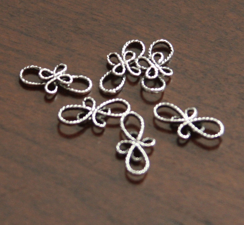 Op de afbeelding: Zes zilveren metalen decoratieve versieringen in de vorm van een gestileerde bloem met een lusontwerp.