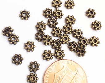 Pode incluir: Um close-up de um monte de pequenas contas de metal douradas. As contas têm a forma de uma flor com um pequeno furo no centro. Um centavo de euro é incluído para escala.