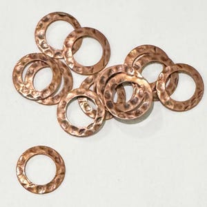 May include: A group of 11 copper jump rings with a hammered texture. The rings are all the same size and are arranged in a loose pile.