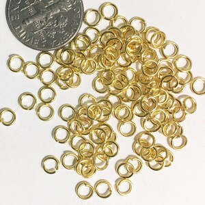 May include: A large pile of gold jump rings, scattered on a white surface. The rings are small and round, with a smooth finish. A 10 pence coin is included for scale.
