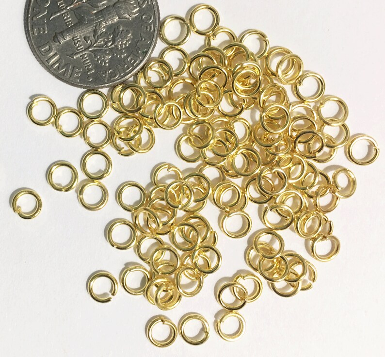 May include: A large pile of gold jump rings, scattered on a white surface. The rings are small and round, with a smooth finish. A 10 pence coin is included for scale.