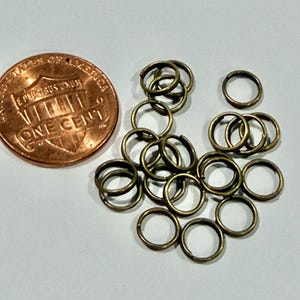 May include: A collection of small, antique brass-colored jump rings scattered on a white surface. A copper one-cent coin is next to the rings for size comparison. The rings are circular and appear to be made of metal.