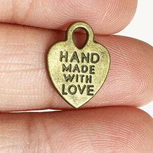 May include: A gold-coloured metal heart charm with the text "HAND MADE WITH LOVE" engraved on it. The charm has a loop at the top for attaching to a necklace or bracelet.