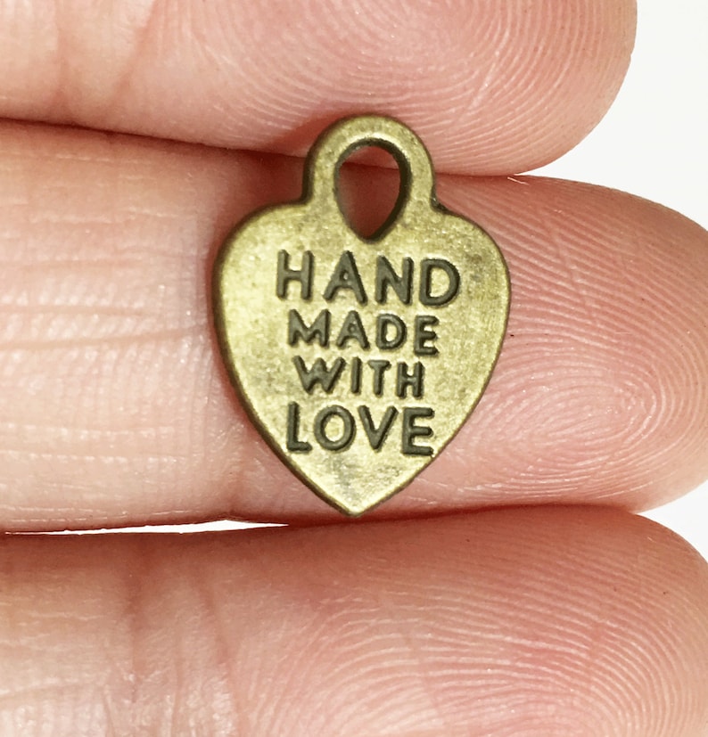 May include: A gold-coloured metal heart charm with the text "HAND MADE WITH LOVE" engraved on it. The charm has a loop at the top for attaching to a necklace or bracelet.