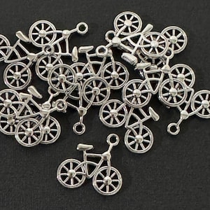 May include: A collection of silver-toned metal bicycle charms, each featuring a detailed frame, two spoked wheels, a seat, and handlebars. These small, flat charms include a circular loop at the top for attachment, arranged in a scattered pile.