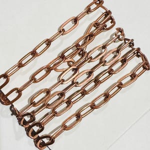 Puede incluir: Primer plano de varias hebras de eslabones de cadena de color cobre. Los eslabones tienen forma ovalada y están conectados en un patrón lineal. Las cadenas están dispuestas diagonalmente sobre una superficie blanca, creando una apariencia texturizada y metálica.