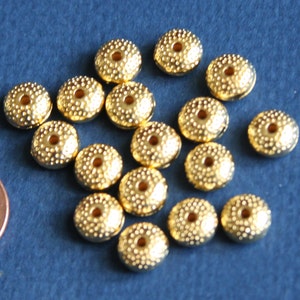 May include: A collection of 17 gold-colored metal beads with a textured surface and a hole in the center. The beads are arranged in a random pattern on a blue surface.