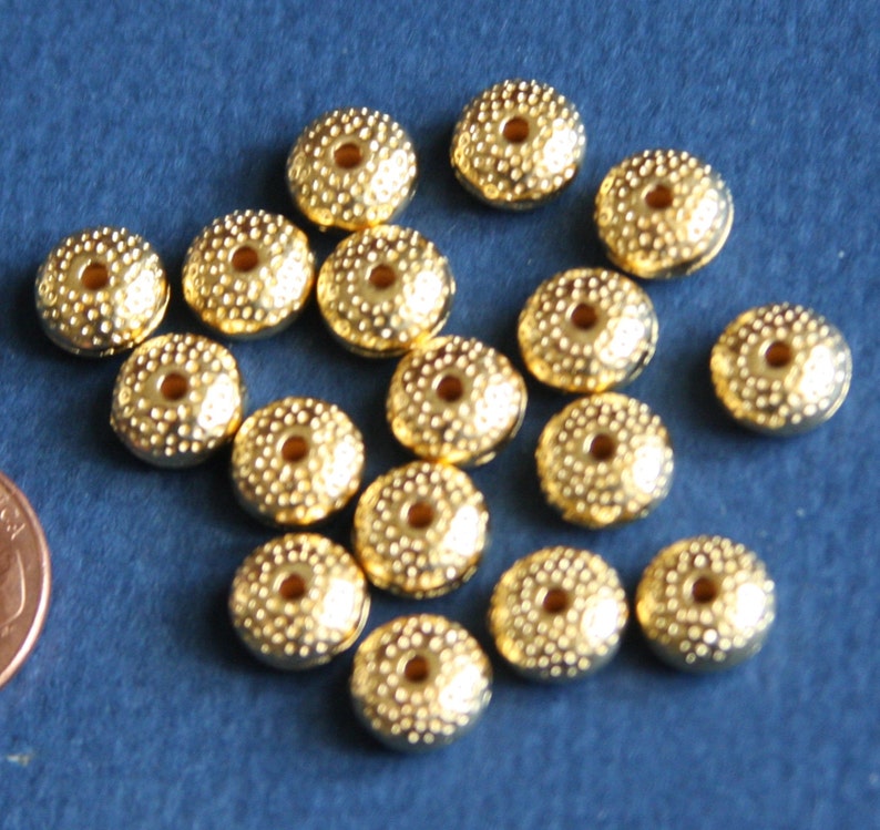 May include: A collection of 17 gold-colored metal beads with a textured surface and a hole in the center. The beads are arranged in a random pattern on a blue surface.