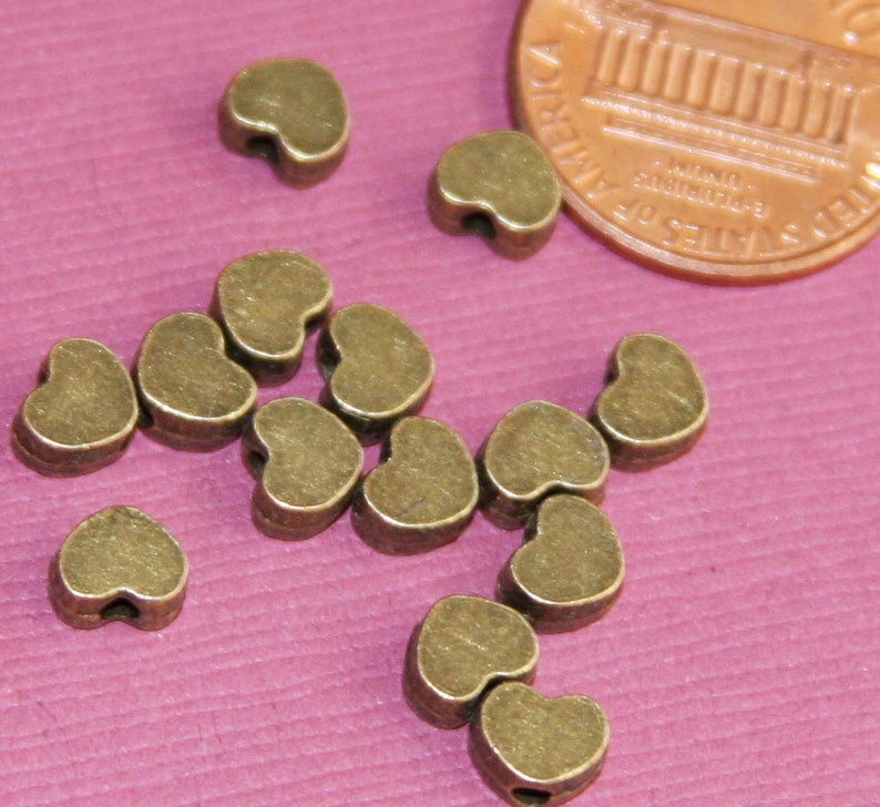May include: A group of 12 antique gold heart-shaped beads with a hole through the centre. The beads are scattered on a pink surface.