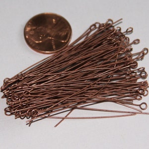 May include: A pile of copper-colored head pins with a loop at the end. The pins are about 1 inch long and are next to a US penny for size comparison.