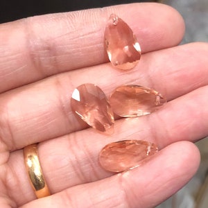 May include: Four teardrop-shaped crystal pendants in a peachy-pink color. The crystals are faceted and have a slight shimmer.