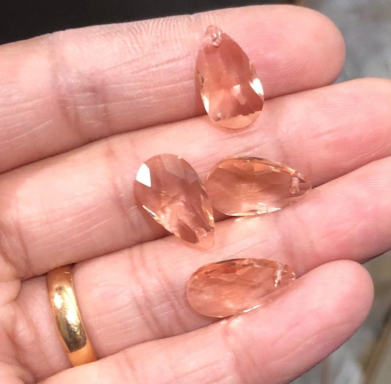 May include: Four teardrop-shaped crystal pendants in a peachy-pink color. The crystals are faceted and have a slight shimmer.