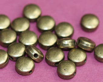 May include: A pile of round, metallic, gold-colored beads with a hole in the center. The beads are scattered on a pink surface.