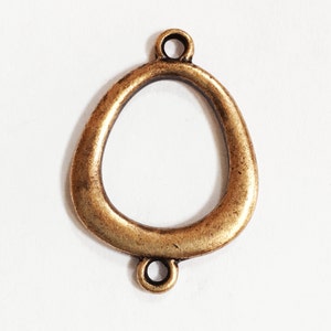 May include: A gold-toned metal pendant with a large oval opening and two smaller loops at the top and bottom. The pendant is designed for attaching to jewelry or other crafts.