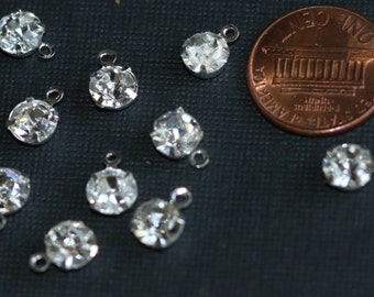 May include: A close-up of ten clear crystal rhinestones with silver settings and loops. The rhinestones are arranged on a dark gray surface. A penny is included for size reference.