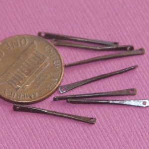 以下が含まれることがあります： 8 本の小さな金属製の編み針のクローズアップ。編み針はピンクの表面に一列に並んでいます。スケールのために、米国の1セント硬貨が背景にあります。