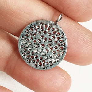May include: A silver-colored, round pendant with a detailed openwork design. The pendant has a small loop at the top for attaching to a chain. The intricate pattern creates a lace-like effect, suitable for jewelry making or crafting.