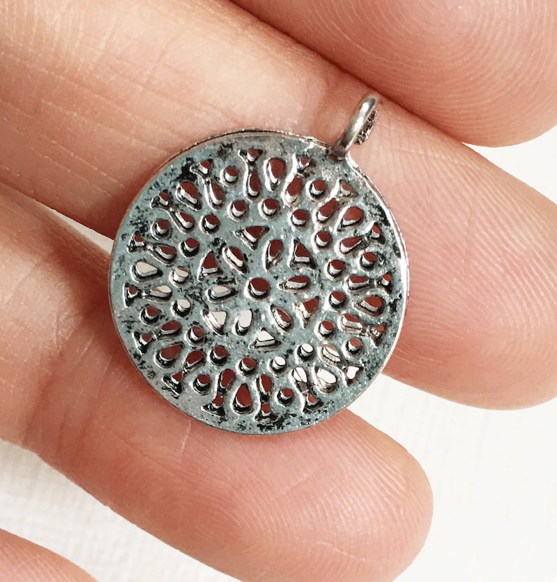 May include: A silver-colored, round pendant with a detailed openwork design. The pendant has a small loop at the top for attaching to a chain. The intricate pattern creates a lace-like effect, suitable for jewelry making or crafting.