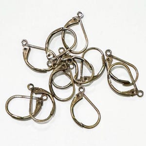 May include: A group of gold-toned earring hooks, also known as ear wires, are piled together. The hooks are made of metal and have a teardrop shape.