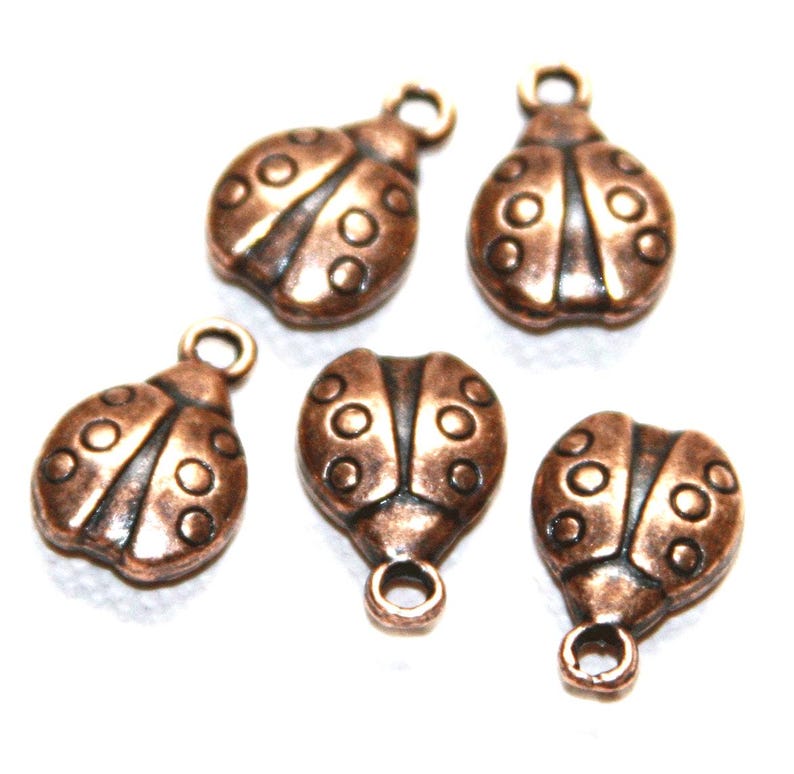 May include: Five small ladybug-shaped charms made of a metallic copper-toned material. Each charm features a rounded body with embossed circular spots and a single circular loop at the top for attachment, arranged in a scattered formation.