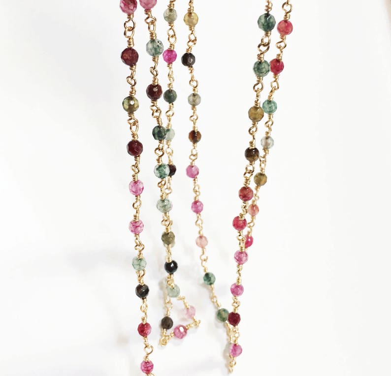 May include: A gold chain necklace with multi-colored gemstone beads. The beads are a mix of pink, green, brown, and black.
