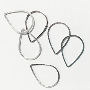 May include: Five silver teardrop-shaped wire hoops, some overlapping, on a white background.