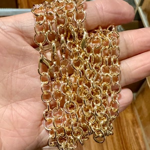 May include: Gold-colored chain with a textured, open-link design. The chain is composed of oval-shaped links and features several lobster claw clasps. The chain is likely for a purse or necklace.