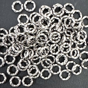 May include: A collection of numerous small, silver-toned metal jump rings, each featuring a distinct twisted or rope-like texture along the circular band. These individual rings are scattered in a dense pile, showing a consistent metallic finish.