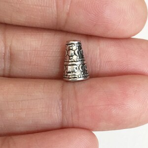 May include: Silver metal bead cap with an intricate design. The bead cap is cone-shaped and has a textured surface.