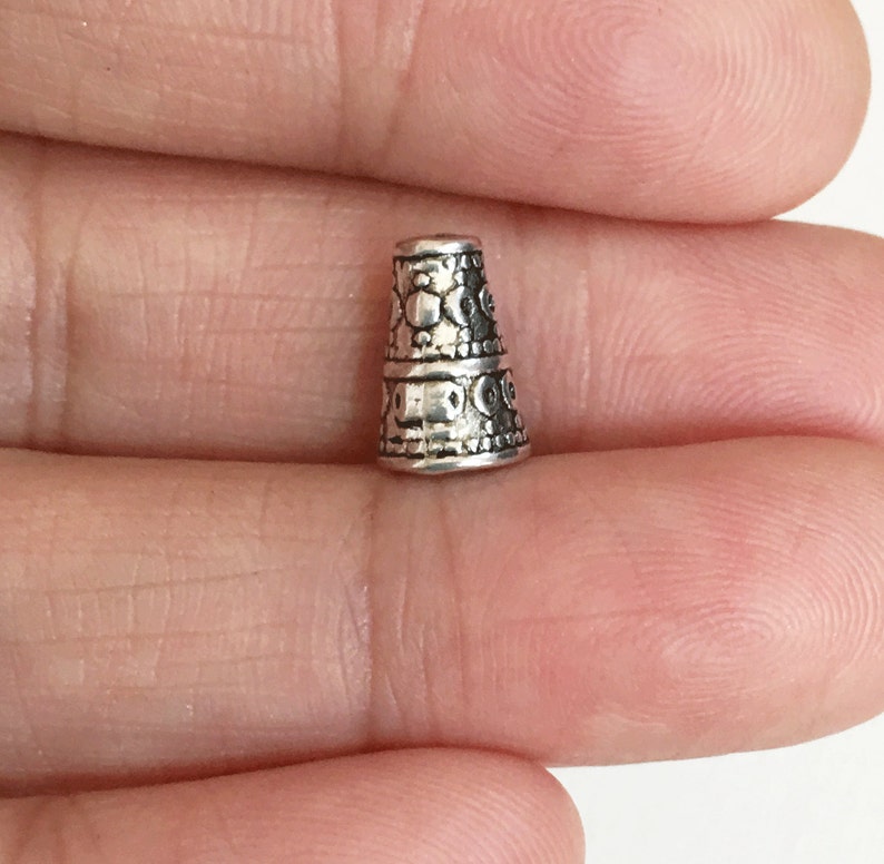May include: Silver metal bead cap with an intricate design. The bead cap is cone-shaped and has a textured surface.