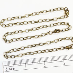 May include: Three gold-toned metal chains of varying lengths, laid out on a white surface with a ruler marked in inches. The chains are made of oval links.