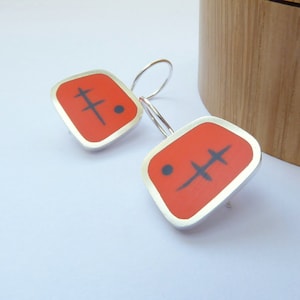Peut inclure: Une paire de boucles d'oreilles carrées en argent et rouge avec un motif abstrait noir. Les boucles d'oreilles sont de forme carrée avec une bordure argentée.