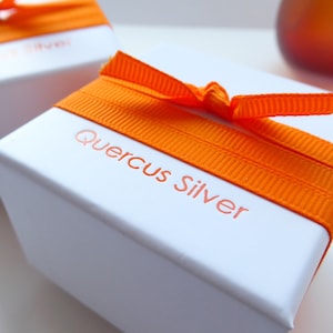 A white jewellery box wrapped with orange ribbon with the name Quercus Silver printed in orange