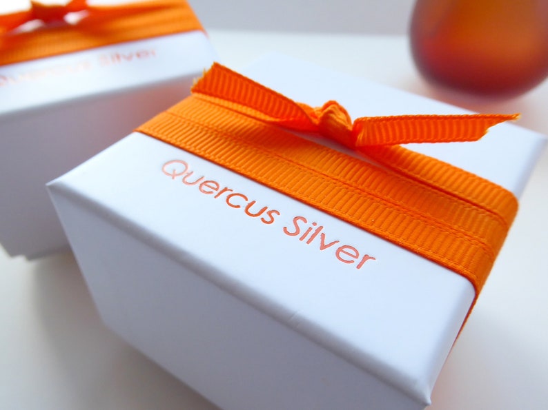 A white jewellery box wrapped with orange ribbon with the name Quercus Silver printed in orange