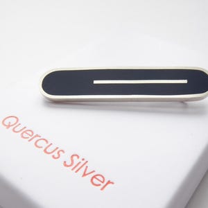 May include: An oval-shaped silver barrette with a black enamel centre and a white stripe. The barrette is displayed on a white box with the words "Quercus Silver" printed in red.