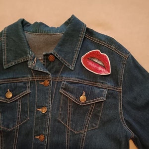 May include: A dark blue denim jacket with a red glitter lips patch on the left side of the chest.