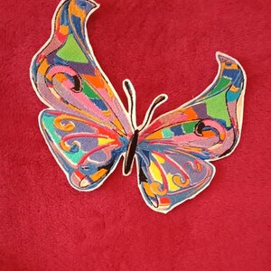 May include: A colourful butterfly embroidered patch with a variety of colours, including blue, green, yellow, orange, pink, and purple. The butterfly is on a red background.