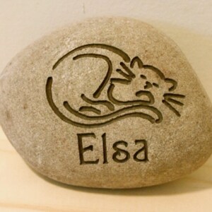 May include: A light brown stone with a carved image of a cat sleeping and the name "Elsa" engraved below.